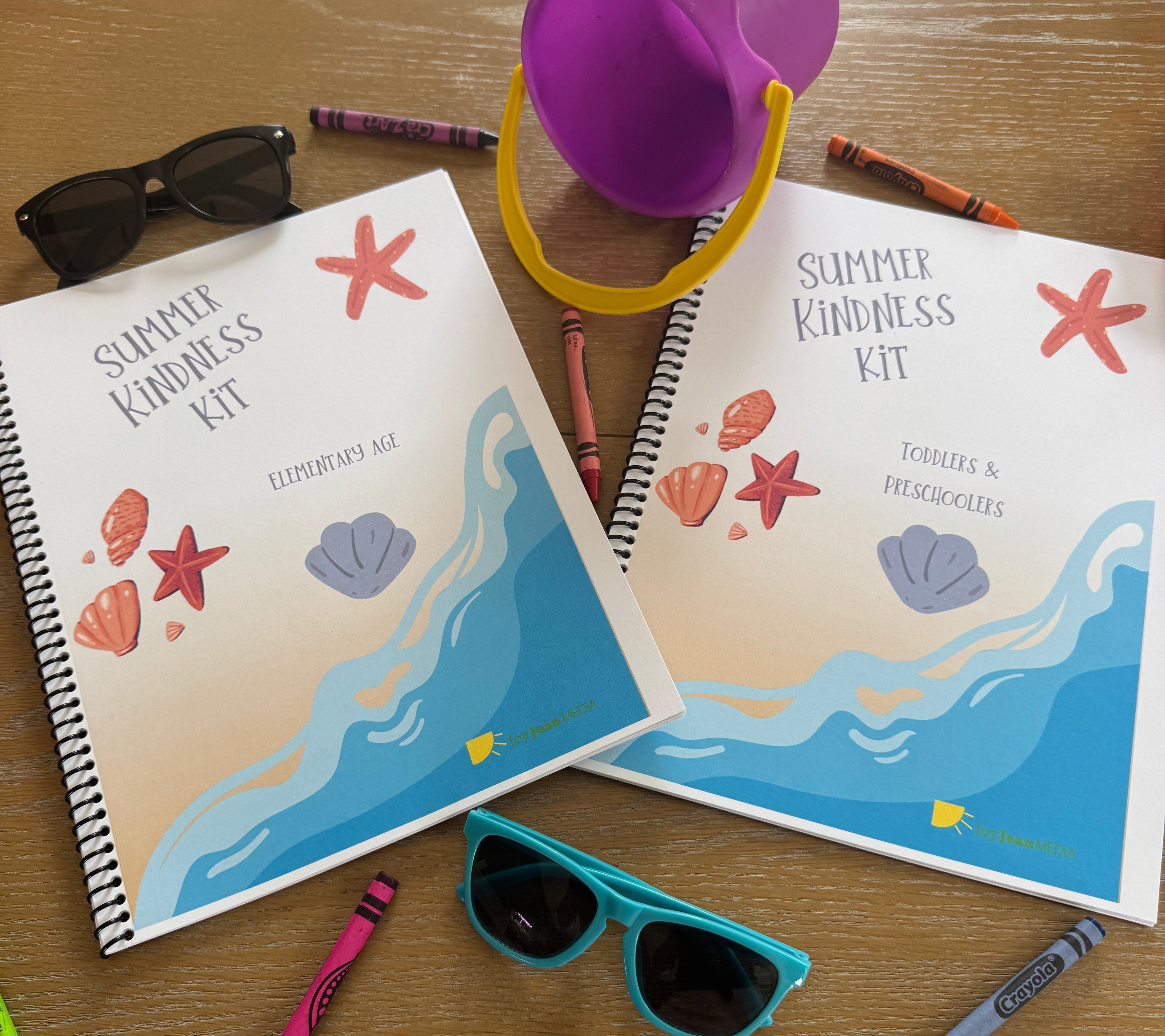 Two spiral-bound notebooks with beach-themed covers, crayons, a purple sand pail, and sunglasses on a wooden surface.